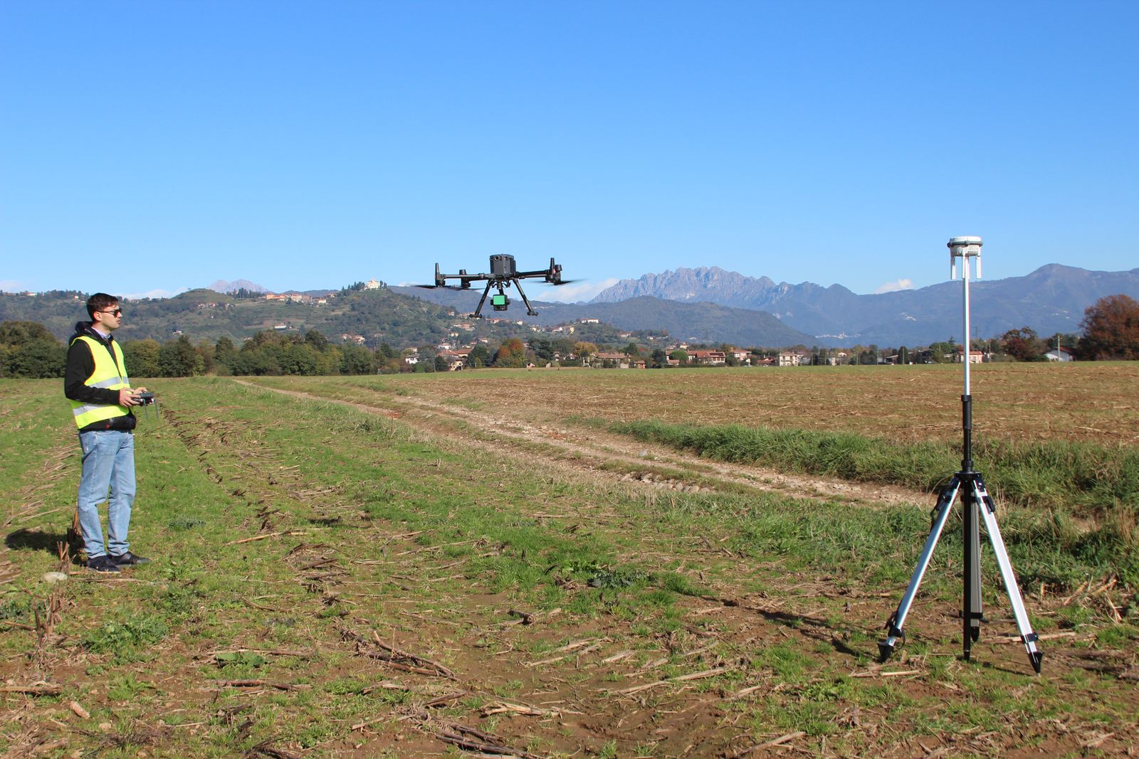 Tecnico Flytech con il radiocomando a sinistra, drone con LiDAR in volo al centro, base GNSS su treppiede a destra, in un campo aperto