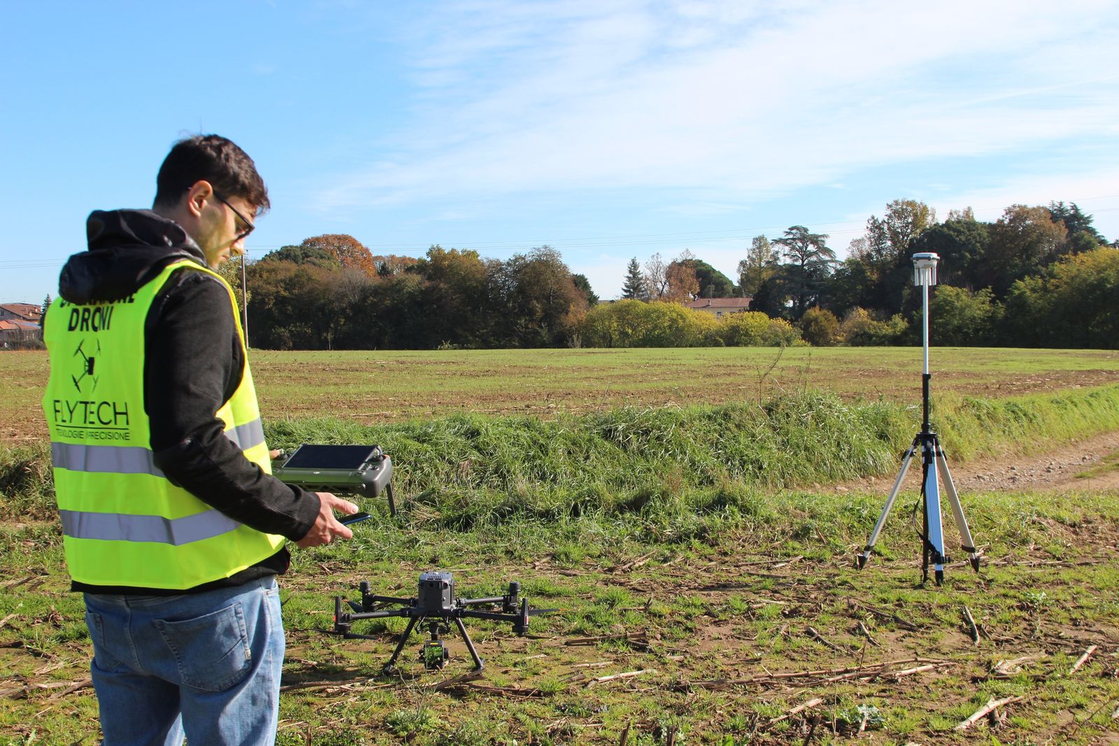 Tecnico Flytech in giubbotto ad alta visibilità con il radiocomando, drone a terra e base GNSS su treppiede in un campo